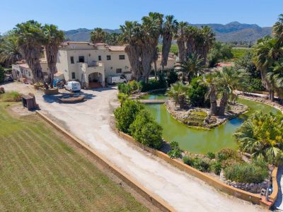 Aerial view of the Mediterranean-style main villa, surrounded by extensive palm trees, a pond with a bridge, and a long driveway, set against a mountain backdrop.