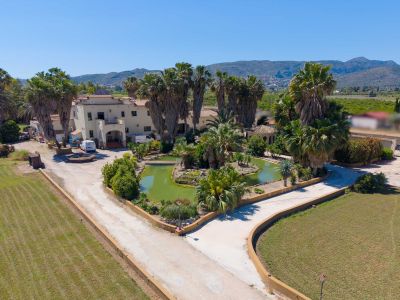 Aerial view showcasing the extensive property with a winding driveway, a large central pond with a bridge, lush palm trees, and the main villa against a mountain backdrop.