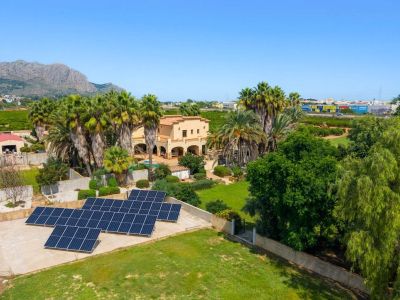 Aerial view showcasing the traditional villa surrounded by palm trees and lush gardens, featuring a prominent solar panel installation and distant mountain vistas under a clear sky.