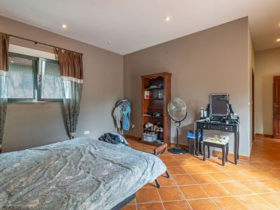 Bedroom featuring a bed, windows with brown and sheer curtains, and terracotta tiled flooring. Includes a black dressing table, wooden bookshelf, and standing fan.