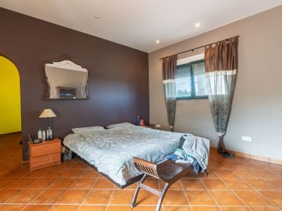 Bedroom featuring a double bed, terracotta tiled floor, and two-tone walls. A window with blinds and curtains provides natural light, complemented by a decorative mirror.