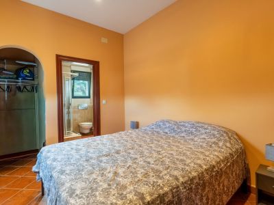 Bedroom featuring warm orange walls and terracotta tiled flooring, furnished with a double bed. An arched opening leads to an open closet, with an en-suite bathroom visible through the door.