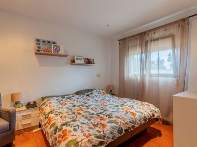 Bright bedroom with a double bed, colorful leaf-patterned duvet, and terracotta tiled floor. Natural light enters through a window with sheer curtains, complemented by a bedside table and wall shelves.