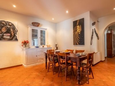 Dining room featuring a dark wooden table with six chairs, terracotta tiled floor, and white walls with recessed lighting. A white hutch and two large paintings adorn the space.
