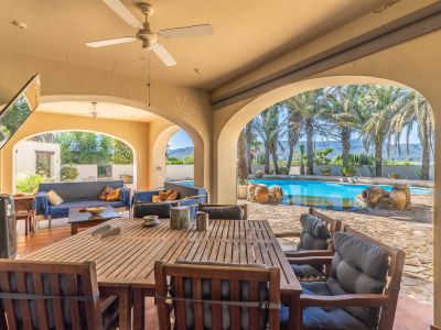 Expansive covered terrace with arches, featuring a wooden dining set and lounge area, offering direct views of the pool, palm trees, and distant mountains.