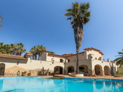 Expansive private swimming pool with clear water, set against a Mediterranean-style villa featuring arched verandas and tall palm trees.