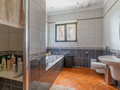 Full bathroom featuring terracotta floor tiles, grey wall tiles with decorative borders, a bathtub, glass-enclosed shower, sink, and a green-framed window.