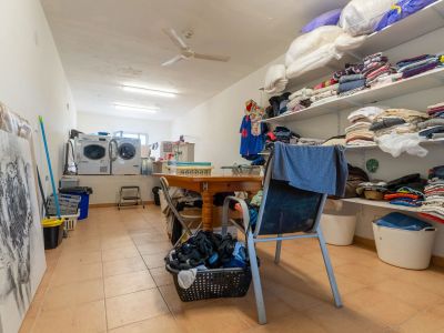 Functional utility room featuring two washing machines, a dryer, and extensive open shelving for laundry and storage. A central wooden table provides a practical workspace, complemented by tiled flooring and ceiling fan.