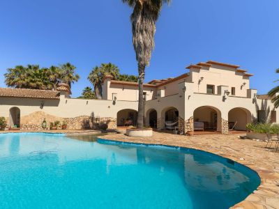 Irregularly shaped private swimming pool with clear blue water, bordered by an expansive natural stone terrace. The Mediterranean-style villa features arched loggias and terracotta roof tiles, amidst mature palm trees.