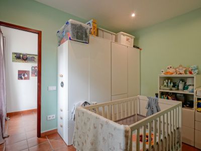 Nursery with light green walls, terracotta tiled floor, and a white crib. Features spacious white wardrobes, a shelving unit, and a changing table, illuminated by a recessed ceiling light.