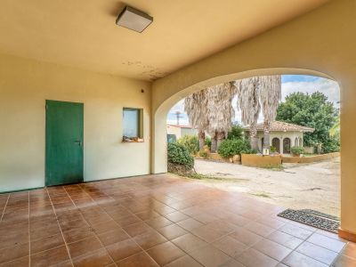 Spacious covered terrace featuring a tiled floor and an archway opening to a gravel path, palm trees, and an exterior building with a tiled roof.