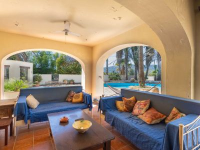 Spacious covered terrace with arches, terracotta flooring, and blue seating, offering views of the pool, palm trees, and distant hills.
