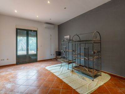 Spacious room with terracotta-tiled floor, two-tone walls, recessed lighting, and an air conditioning unit. Features a double-glazed door with roller shutter and large metal bird cages.