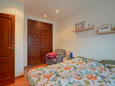 This bedroom features a dark paneled wooden wardrobe and door, terracotta floor tiles, and a bed with a vibrant leaf-patterned duvet. Recessed lighting illuminates the space.