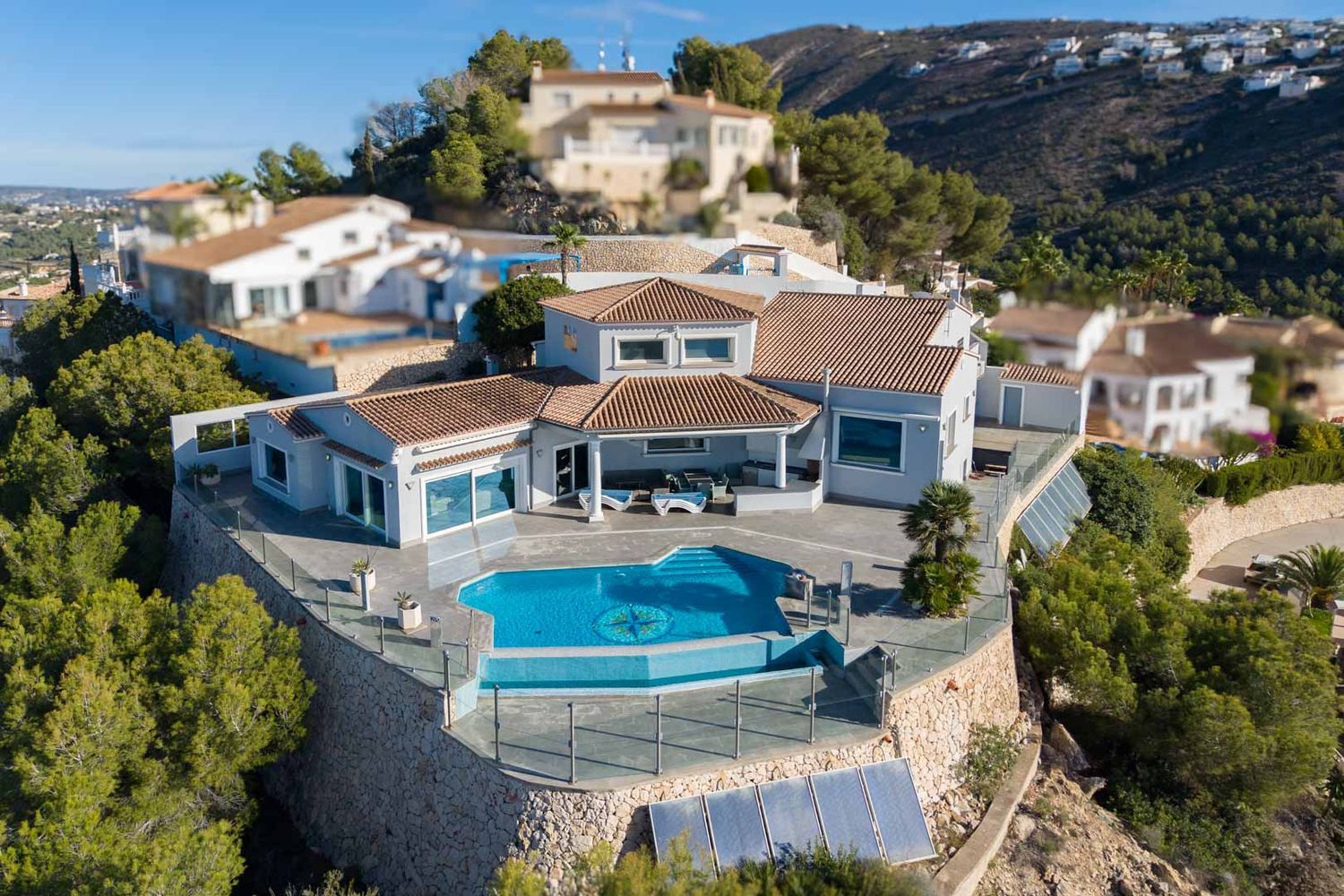 Aerial view of the villa featuring an infinity pool with a compass mosaic and a spacious glass-railed terrace. Terracotta tiled roofs and stone retaining walls define its hillside setting.