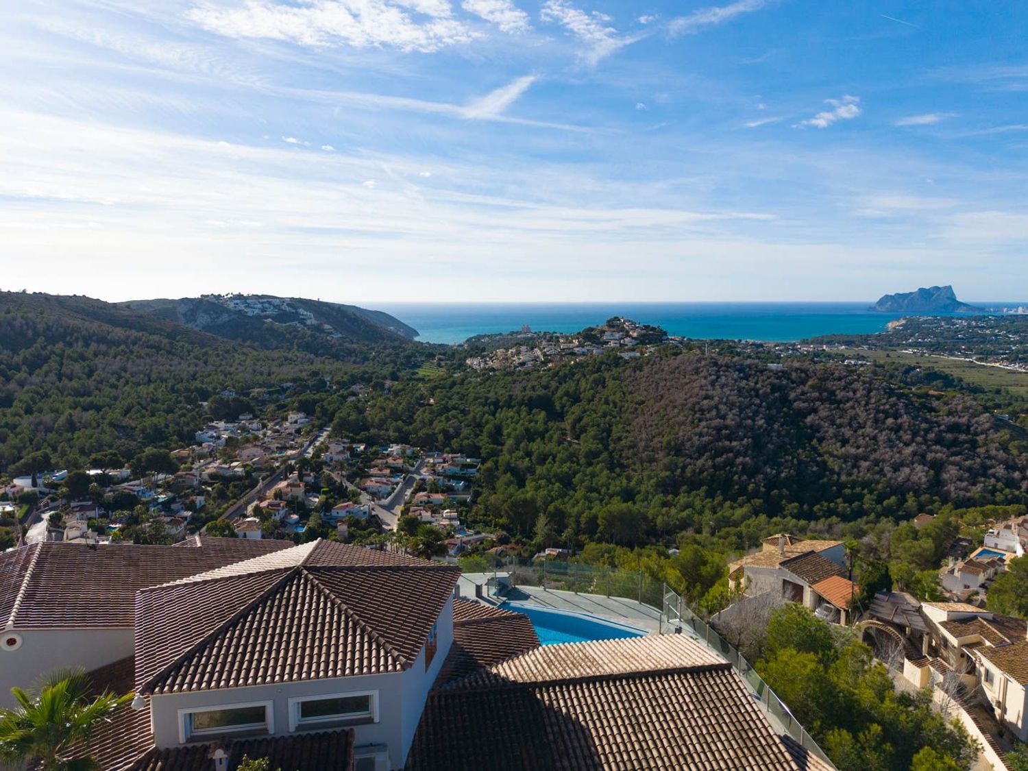 Aerial view showcasing the property's terracotta roofs and pool, nestled amidst green hills, with the Mediterranean Sea and Peñón de Ifach in the distance.