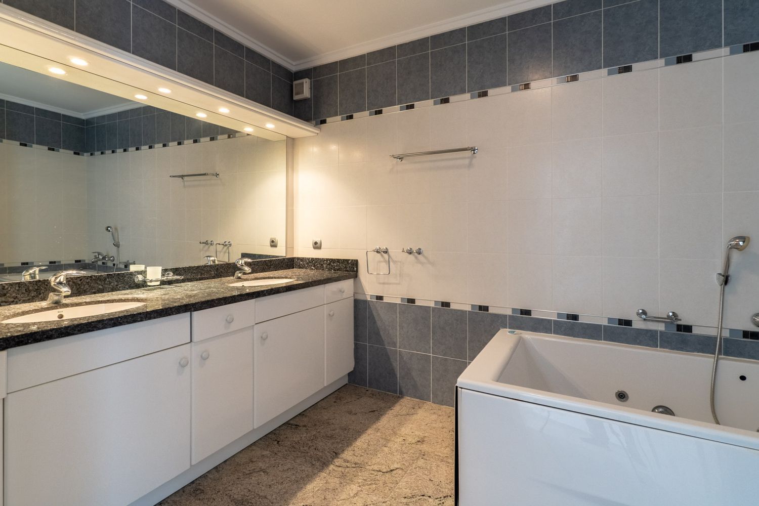 Bathroom featuring a double vanity with dark granite countertop, white cabinets, and an illuminated mirror. Includes a built-in bathtub and grey-white tiled walls.