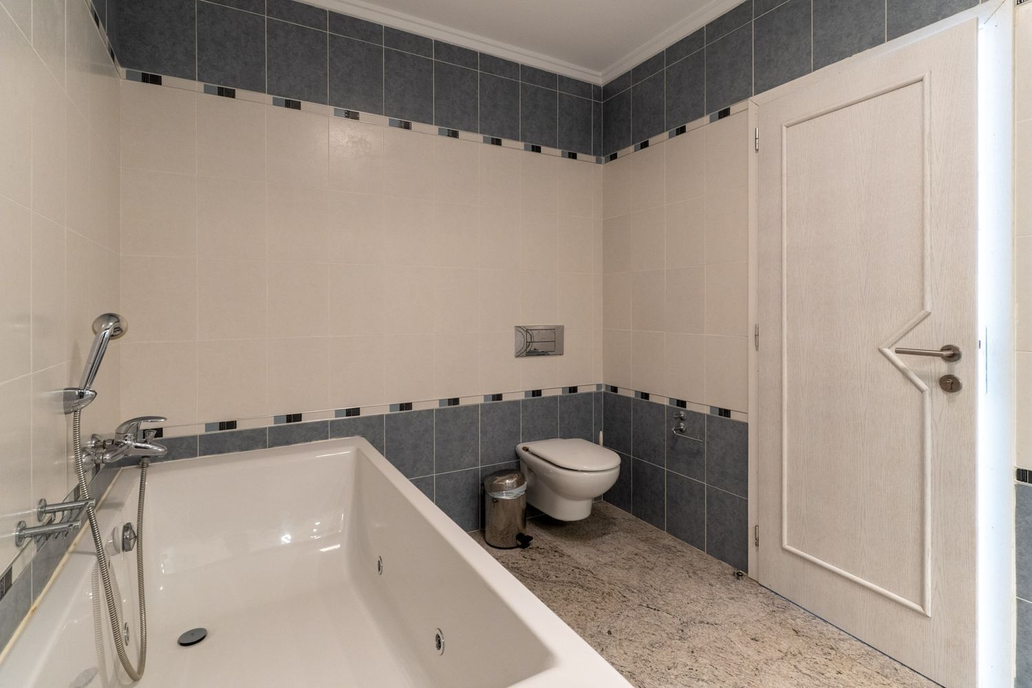Bathroom featuring a white bathtub with integrated shower, beige and grey tiled walls with a decorative border, and light granite flooring. Includes a wall-mounted toilet and a door with a geometric design.