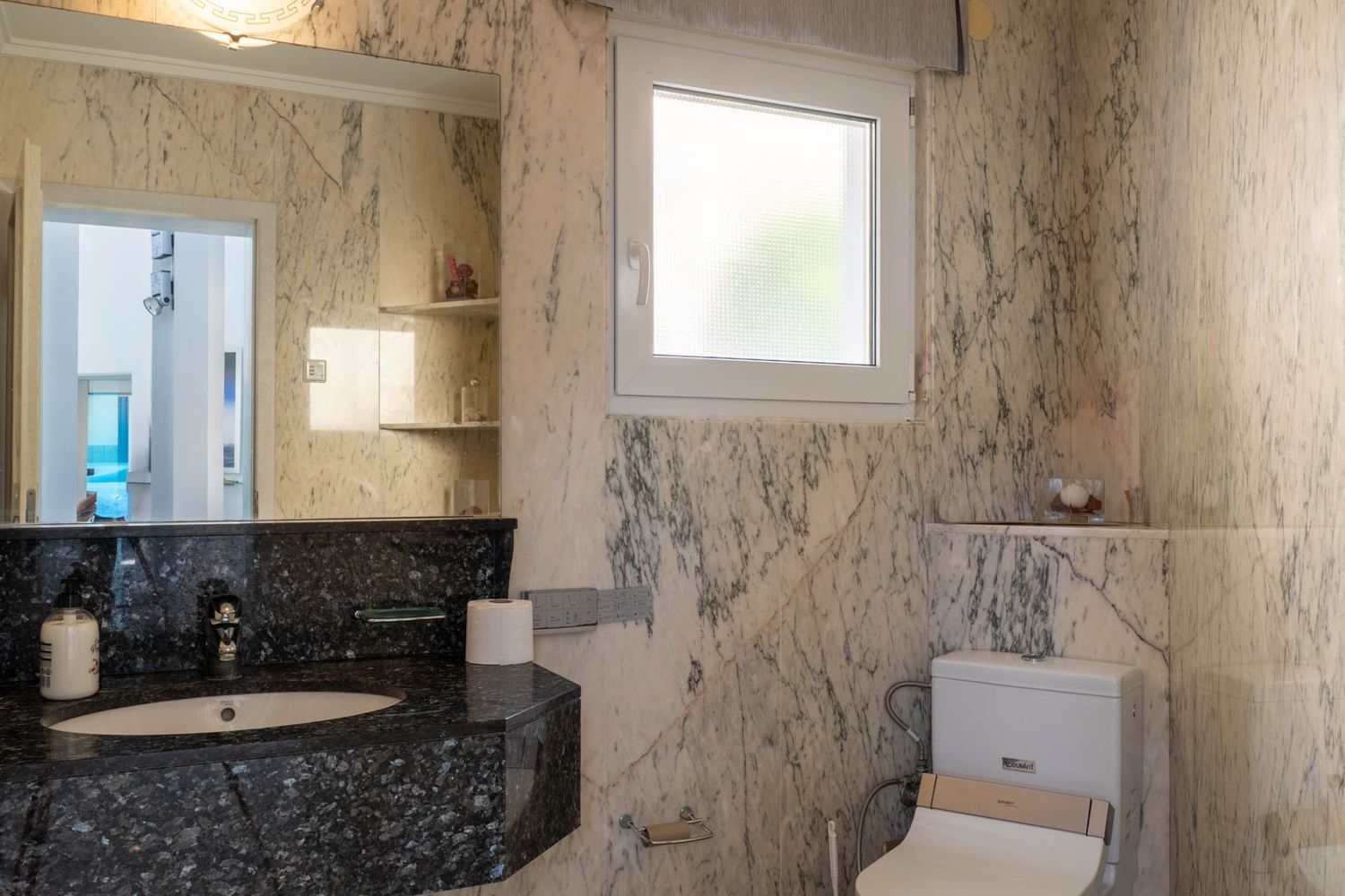 Bathroom featuring light veined marble walls, a dark granite vanity with an integrated sink, and a large mirror. It includes a frosted window and a toilet with bidet functionality.