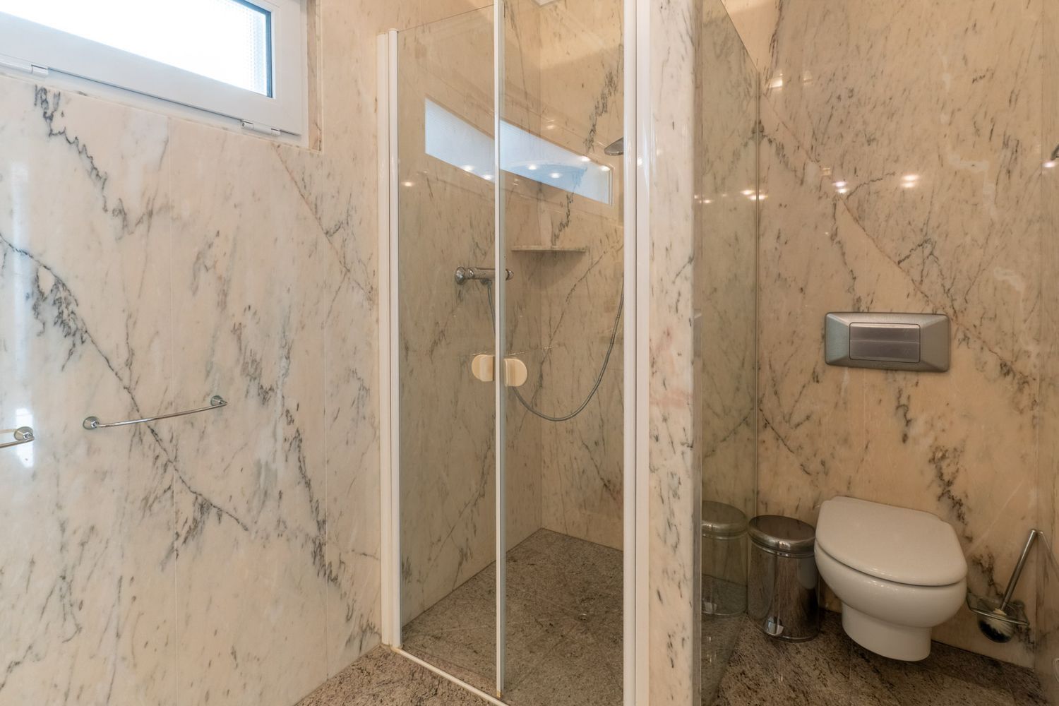 Bathroom featuring veined marble walls and granite flooring. It includes a glass-enclosed shower with an integrated shelf and a wall-mounted toilet, illuminated by a high window.