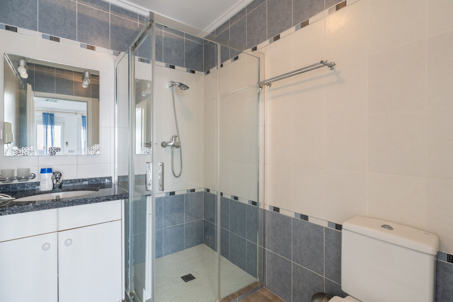 Bathroom with a dark speckled granite vanity top, a decorative mirror, and a glass-enclosed shower. Walls feature light beige and blue-grey tiles with a mosaic border.