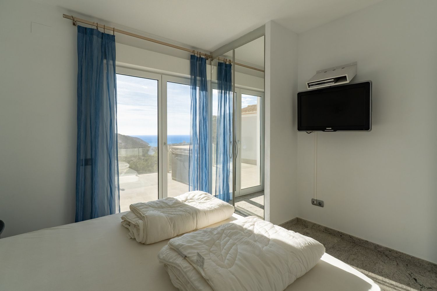 Bedroom featuring a bed with white linen, a wall-mounted TV, and large sliding glass doors with sheer blue curtains, opening to a terrace with panoramic sea views.