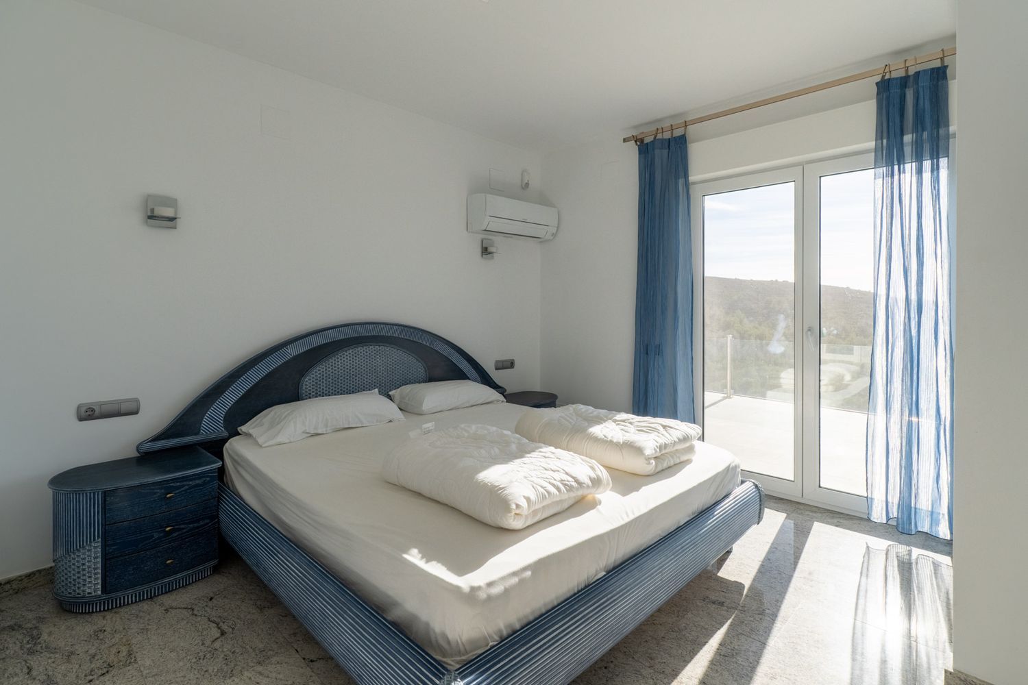 Bedroom featuring a blue patterned bed frame with matching nightstands, light-colored flooring, and direct access to a terrace through a glass door, offering hillside views and natural light.