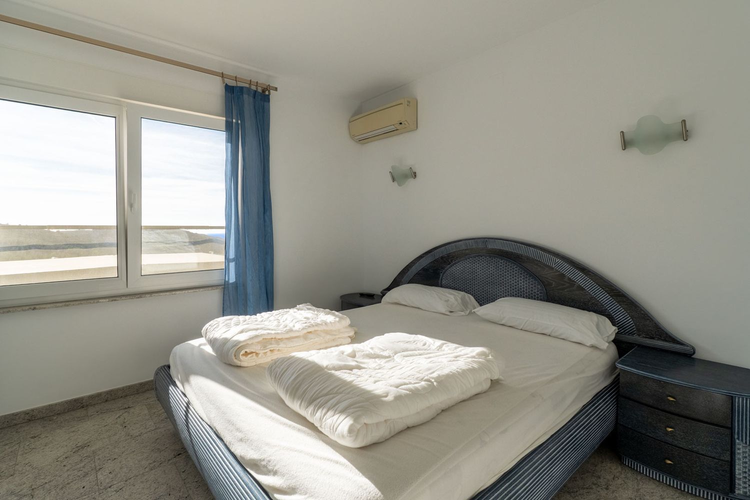 Bedroom featuring a blue patterned bed with a woven headboard, light tiled flooring, and an exterior view through the window. Equipped with air conditioning and a matching bedside table.
