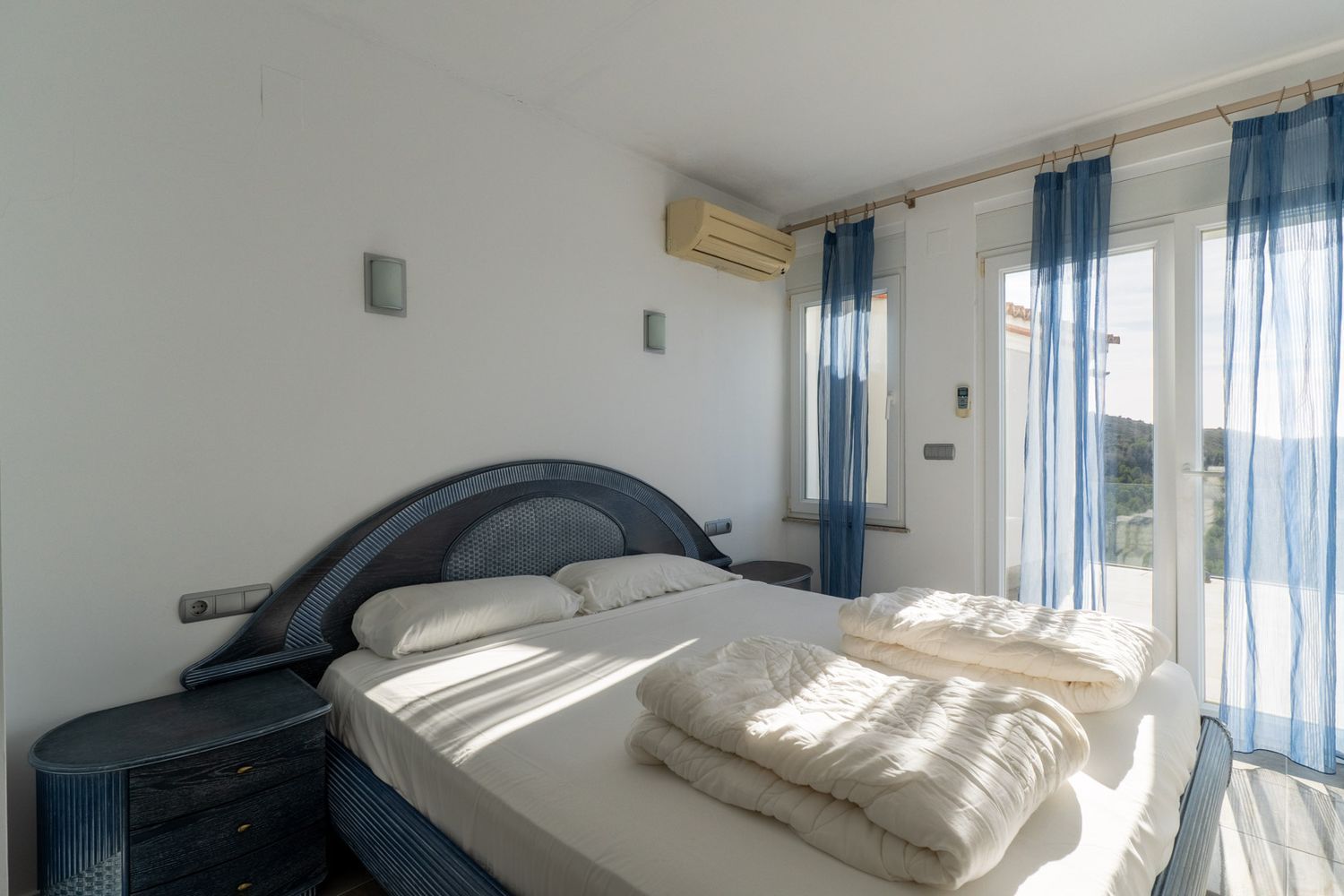 Bedroom with a blue and dark wood double bed, matching bedside tables, and wall lights. Glass doors with sheer blue curtains lead to a terrace, featuring an air conditioning unit.