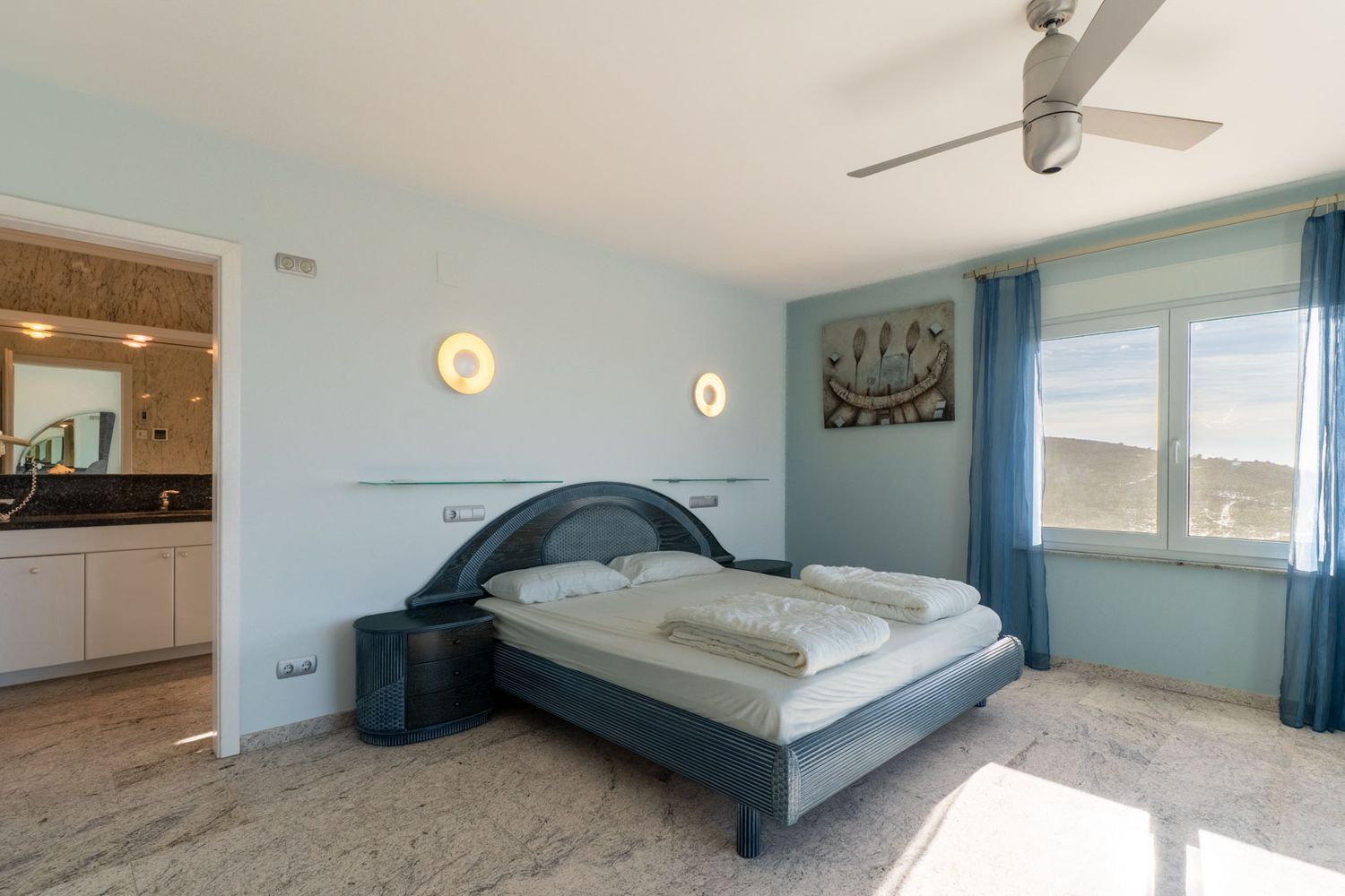 Bedroom with a blue-toned designer bed, matching nightstands, and light marble flooring. The window offers hill views, complemented by blue sheer curtains and an adjacent en-suite bathroom.