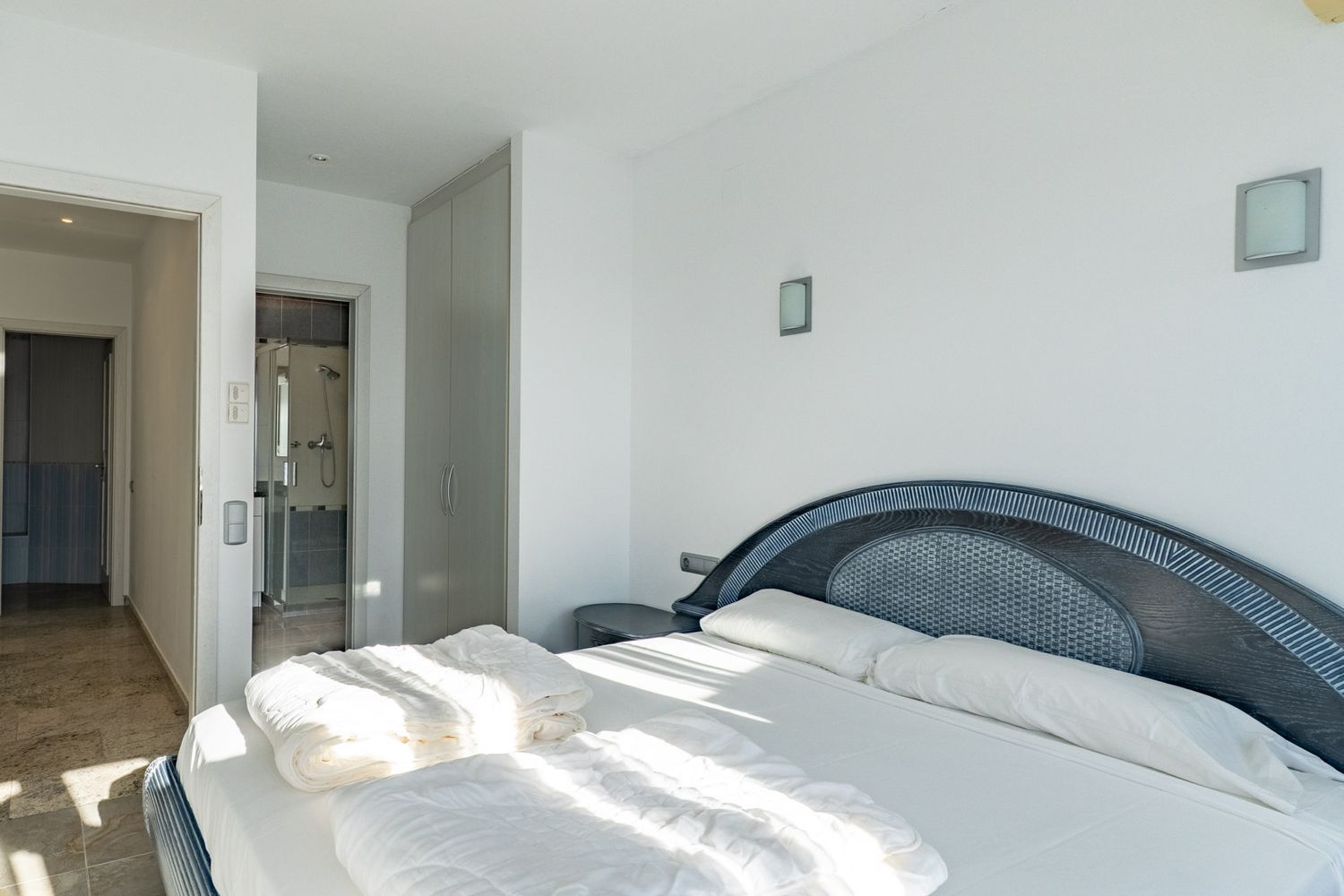 Bright bedroom featuring a double bed with a dark wood headboard and woven inlay. It includes a built-in wardrobe and direct access to an en-suite bathroom with a glass shower.