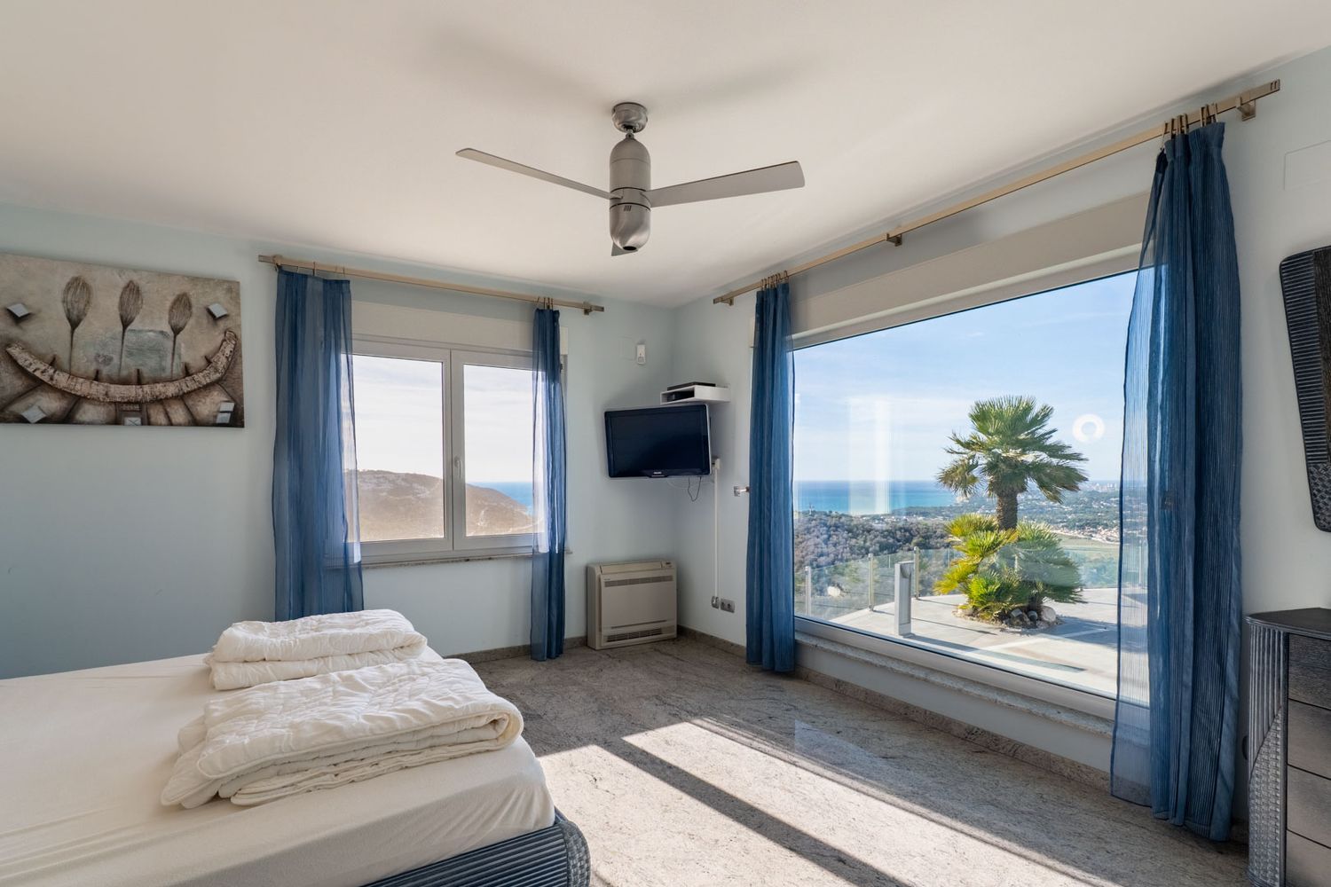 Bright bedroom featuring a large panoramic window with direct sea views and a palm tree. Light granite-effect tiled floor and blue sheer curtains.
