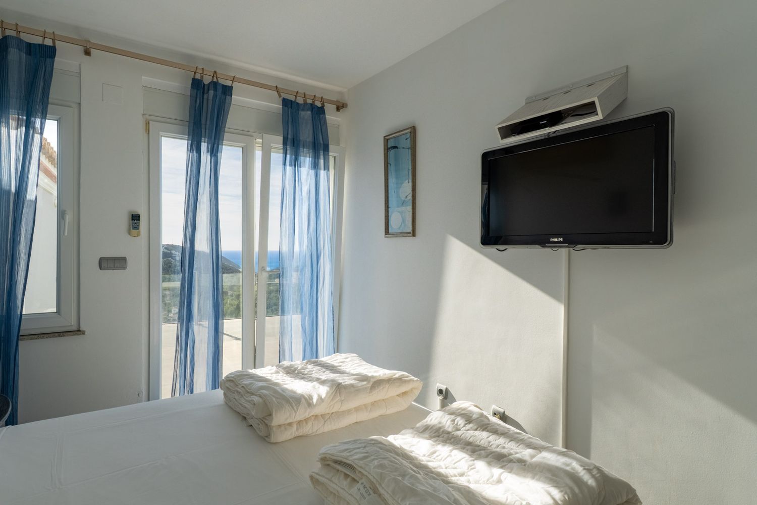 Bright bedroom featuring twin beds and direct terrace access. Sea views are visible through sliding glass doors with sheer blue curtains, complemented by a wall-mounted TV.
