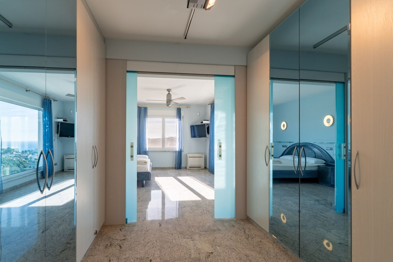 Bright hallway with granite floor and light wood wardrobes. Sliding glass doors frame a bedroom with sea views, while mirrored wardrobe panels reflect another blue-themed bedroom.