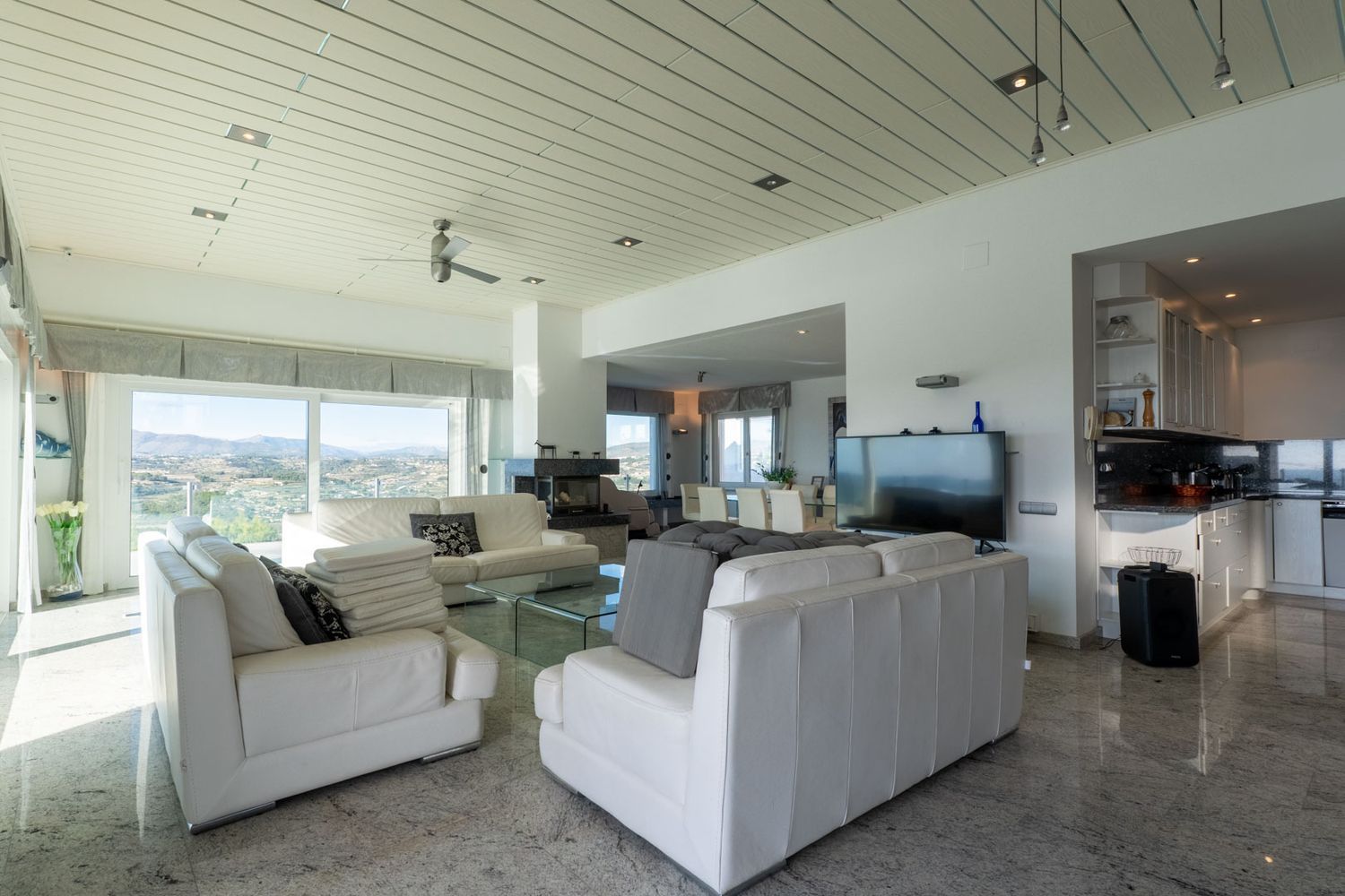 Bright living-dining area with white leather sofas, marble flooring, and large windows offering panoramic mountain views. The open-concept kitchen features dark granite countertops.