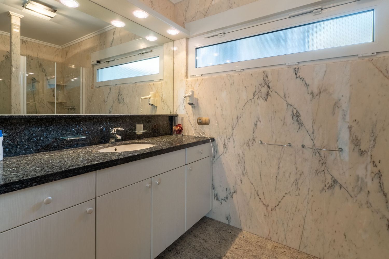 Elegant bathroom featuring veined marble walls and floor, a vanity with a dark granite countertop, and an integrated sink. A frosted horizontal window provides natural light and privacy.