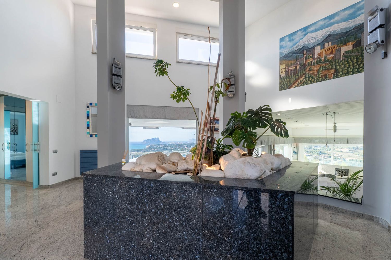 Entrance hall featuring a dark granite decorative island with white stones and plants. High ceilings and large windows frame panoramic sea views, including the iconic Peñón de Ifach.