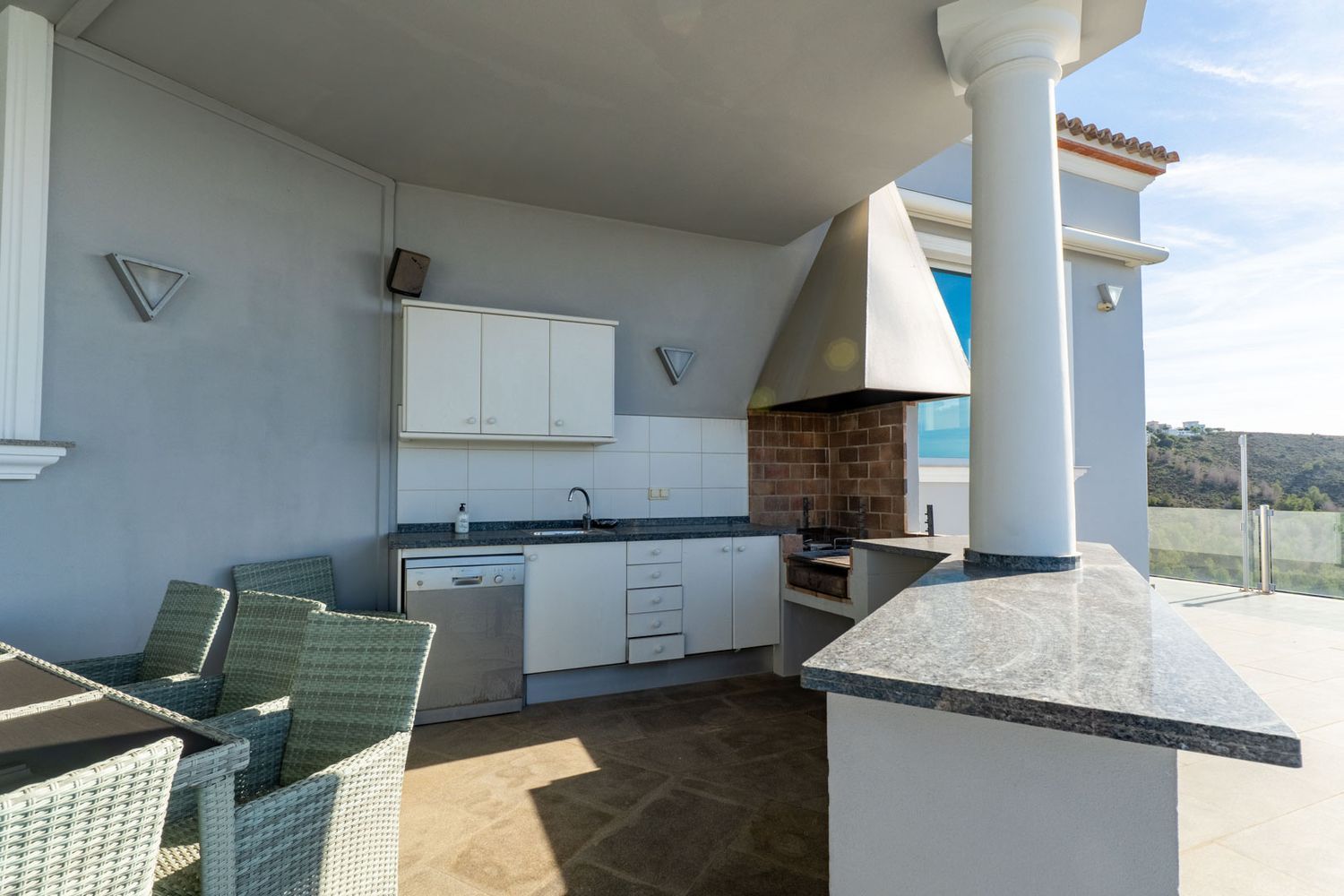 Equipped outdoor kitchen featuring a brick barbecue, granite countertops, sink, and dishwasher, adjacent to a dining table on a covered terrace with hillside views.