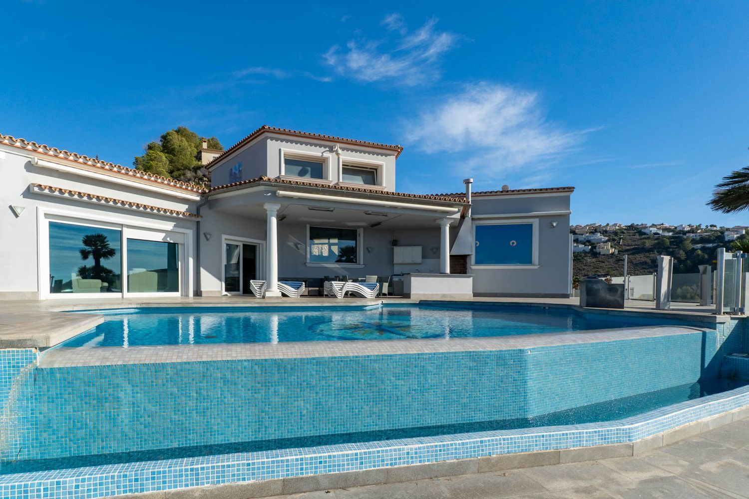 Infinity pool with blue mosaic tiles and covered terrace featuring an outdoor kitchen, adjacent to the villa's facade.