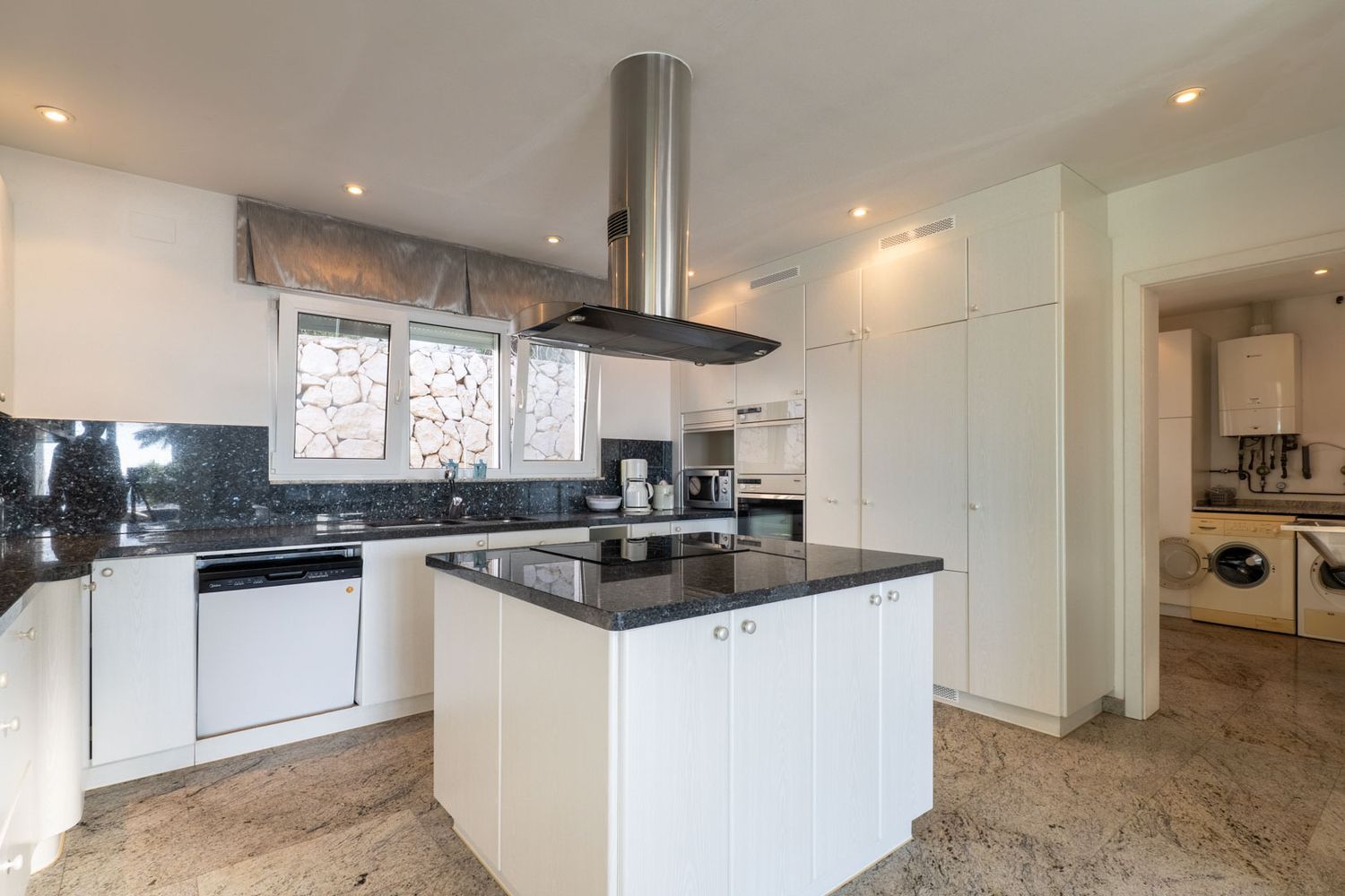 Modern kitchen with white cabinetry, dark granite countertops, and a central island with integrated hob. Integrated appliances, stone wall view from window, and direct access to laundry.