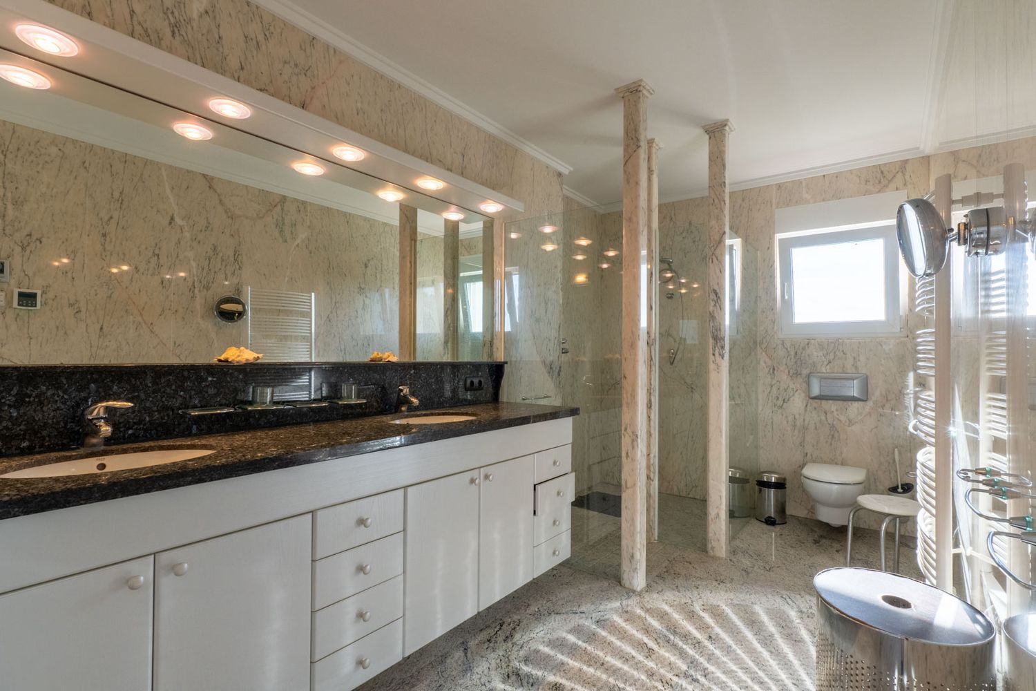 Spacious bathroom featuring a double vanity with dark granite countertop and white cabinetry. The walk-in shower with glass partitions and marble pillars complements the light marble walls and floor.