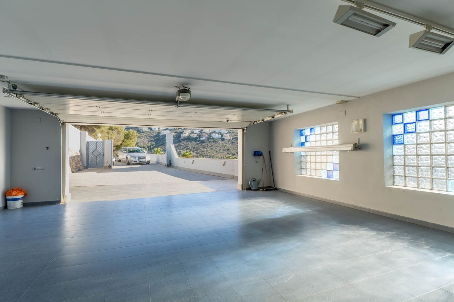 Spacious garage featuring light blue tiled flooring and glass block windows with blue accents, with the main door open to the exterior driveway and distant hills.