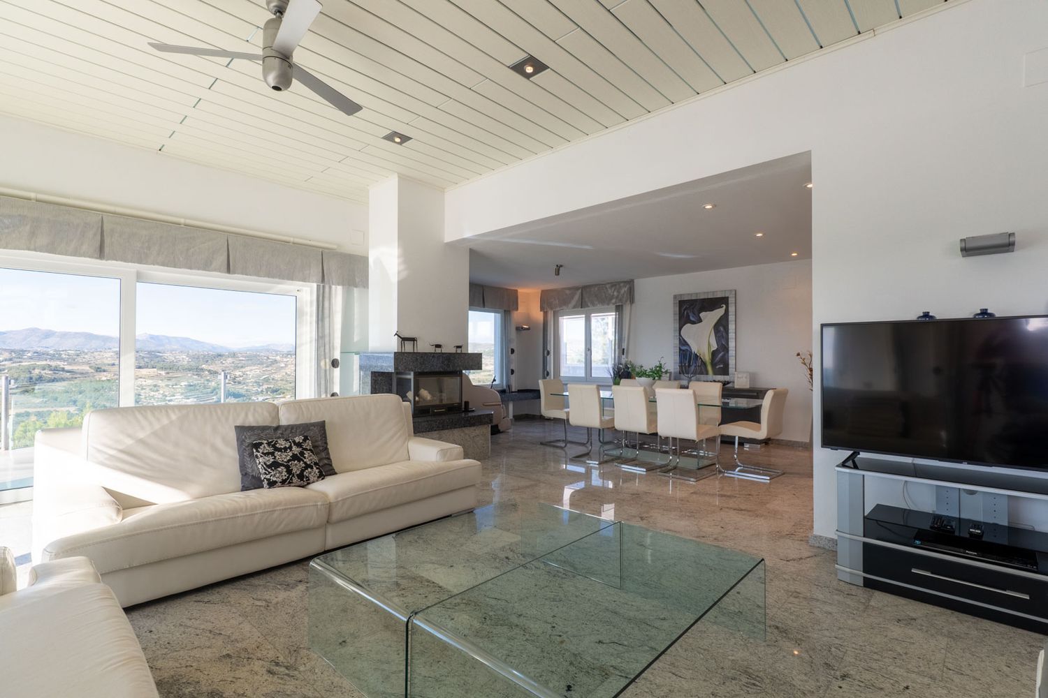 Spacious open-plan living and dining area featuring polished marble floors, a white sofa, and a dark stone fireplace. Large windows frame panoramic mountain and valley views.
