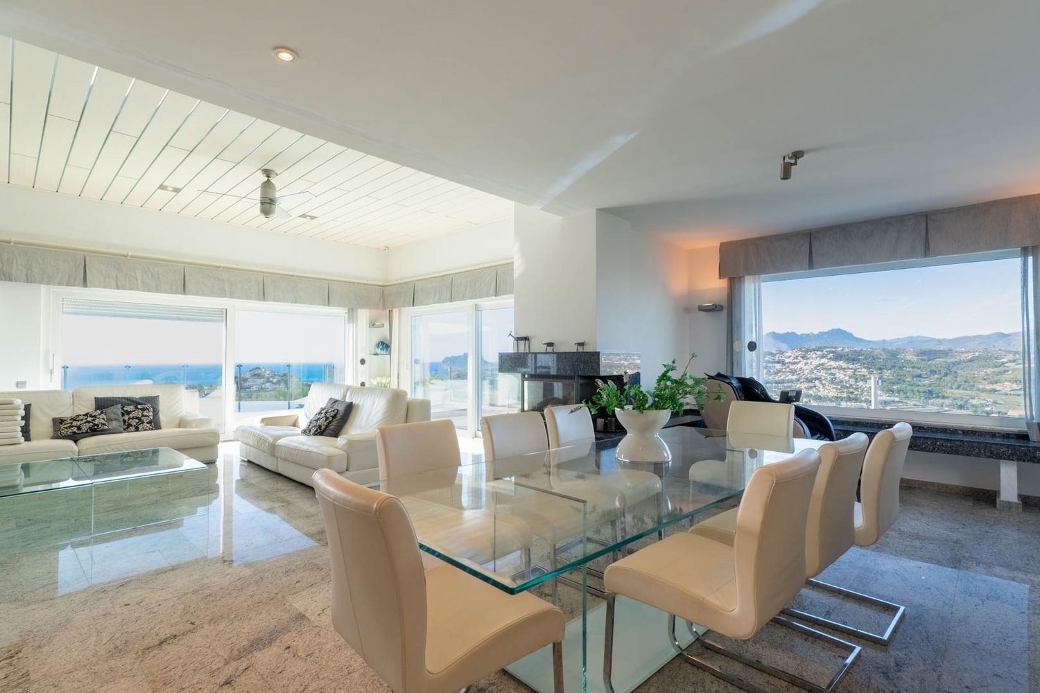 Spacious open-plan living and dining area with polished stone floors. Features a glass dining table, white leather sofas, and a modern fireplace, all bathed in natural light with dual sea and mountain views.