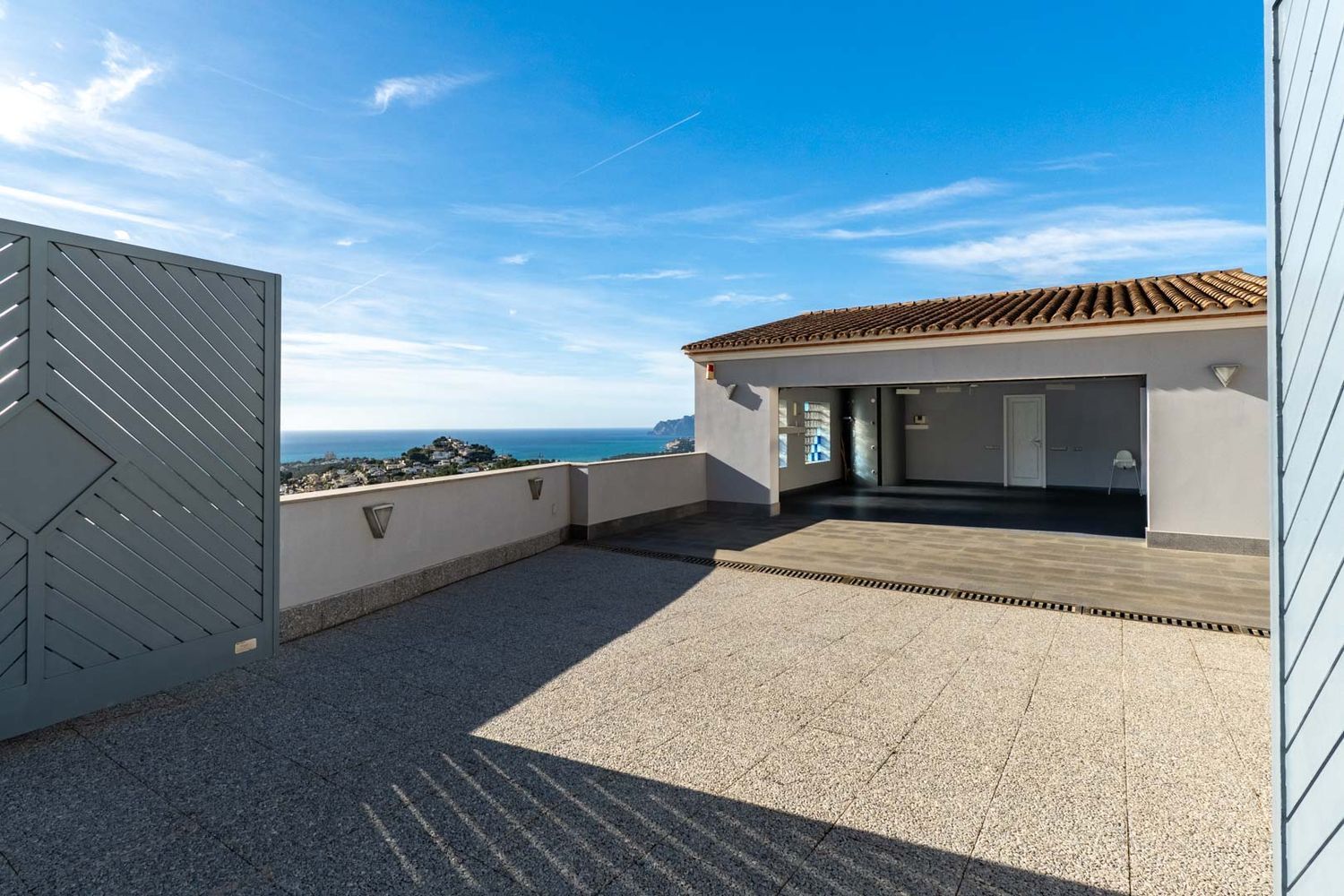 Spacious outdoor area with tiled paving and access to a covered garage. Features panoramic Mediterranean sea and coastal views, framed by a modern grey gate and a low wall with integrated lighting.