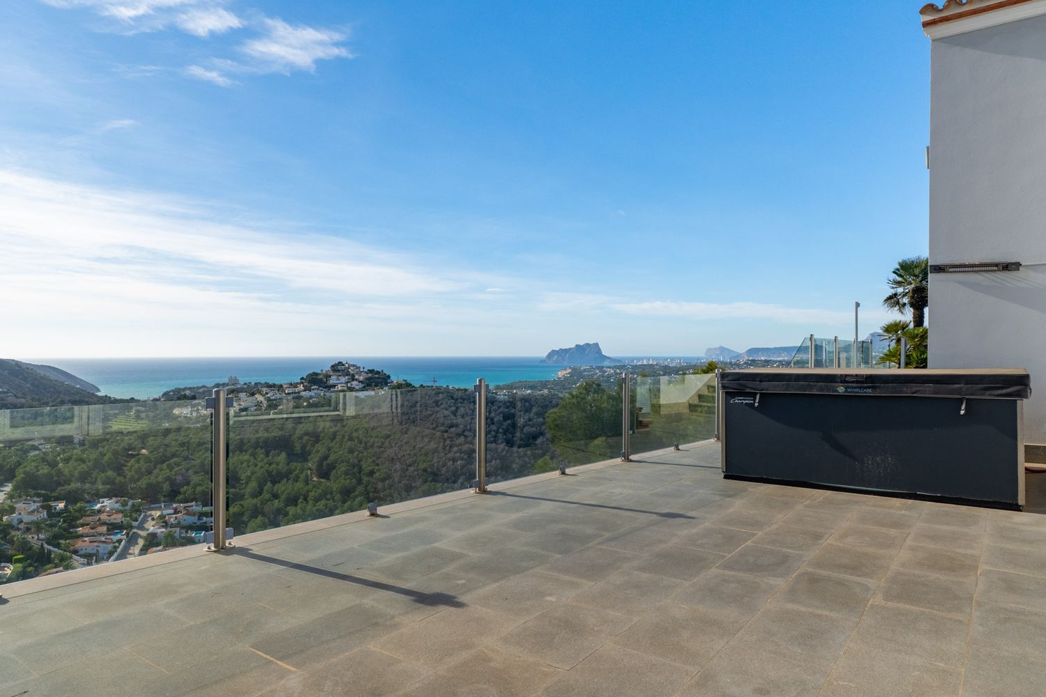 Spacious outdoor terrace with grey tiled flooring and a covered jacuzzi, framed by a glass balustrade showcasing panoramic sea views and the Peñón de Ifach.