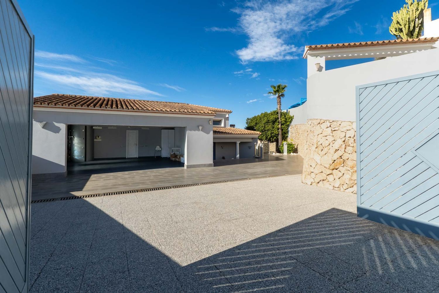 Spacious paved driveway leading to the integrated garage with terracotta tiled roof, framed by a stone wall and a partially open light blue gate under a clear sky.