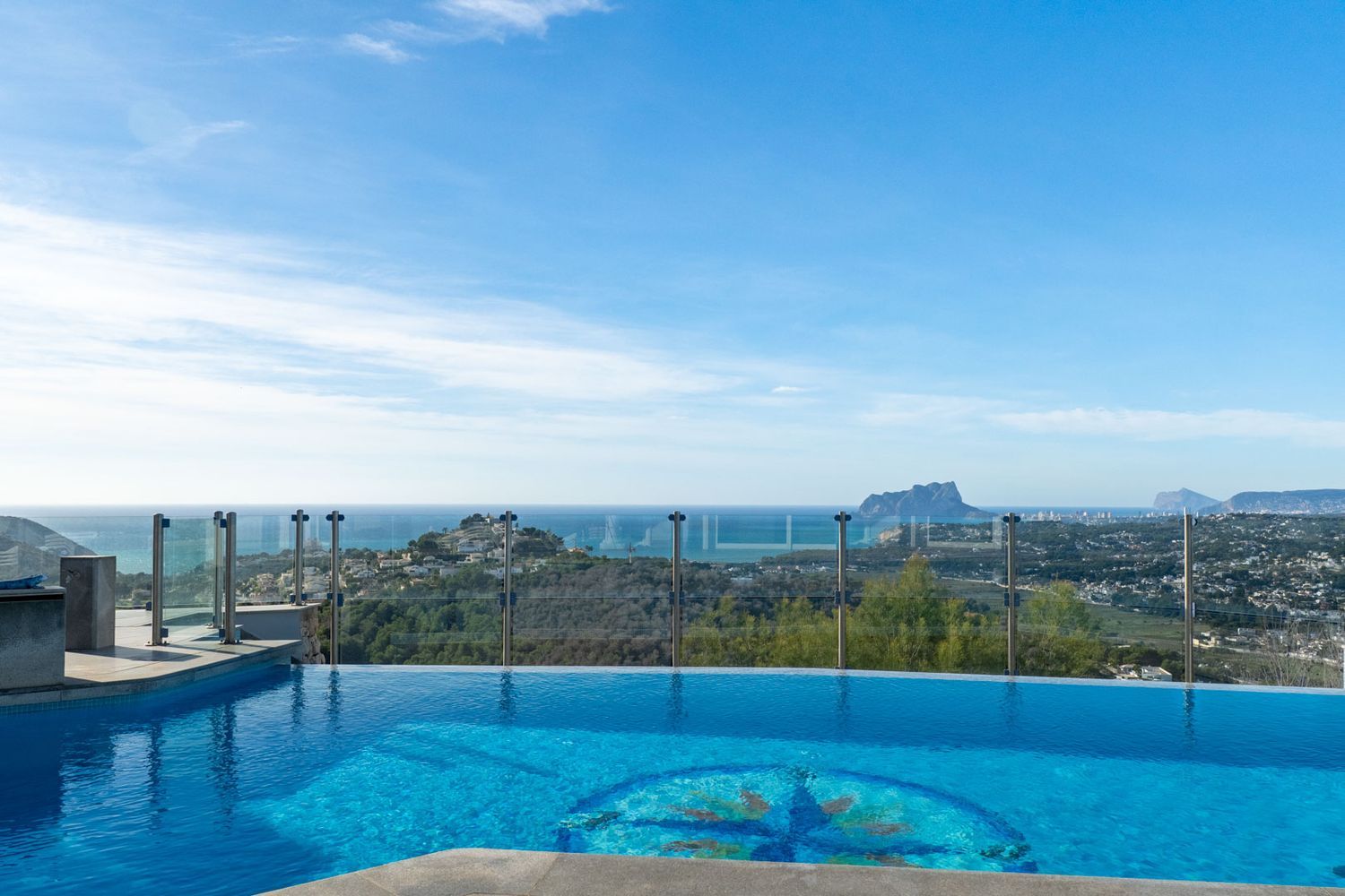 The infinity pool, featuring a distinctive central mosaic, visually merges with the horizon. It offers panoramic sea views, the coastline, and the iconic Peñón de Ifach.