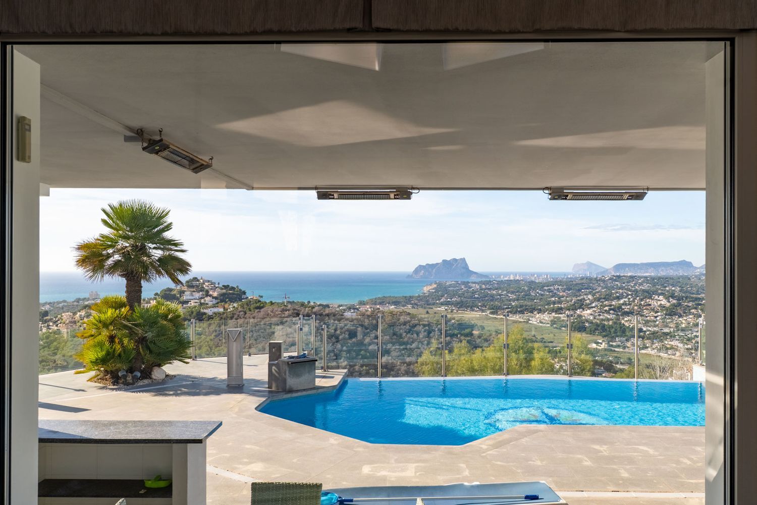 View from inside to the terrace with pool, palm trees, and a vast panorama of the sea, Moraira coastline, and Peñón de Ifach. Ceiling heaters under the overhang.