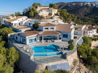 Aerial view of the villa featuring an infinity pool with a compass mosaic and a spacious glass-railed terrace. Terracotta tiled roofs and stone retaining walls define its hillside setting.